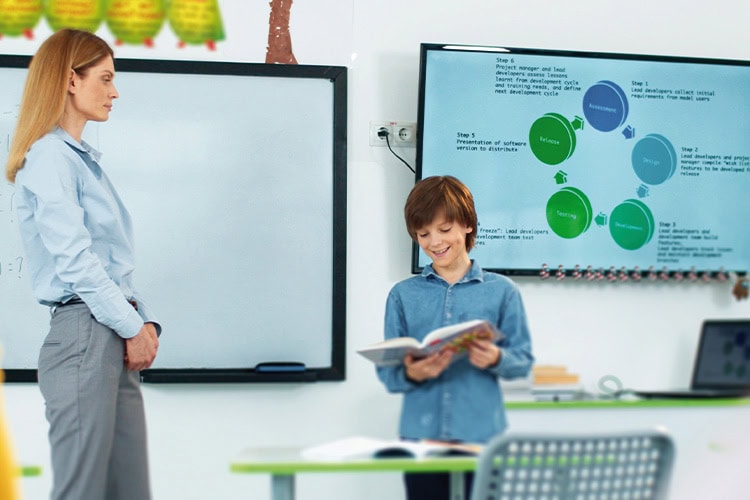 Boy student looking down at a book in his hands while teach is standing on the side of him supervising